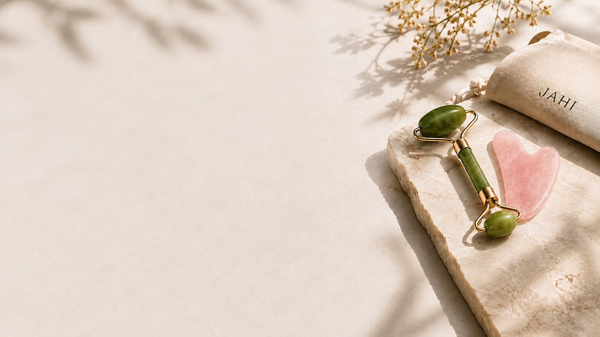 Jade roller and rose quartz gua sha resting on a stone slab beside a Jahi pouch